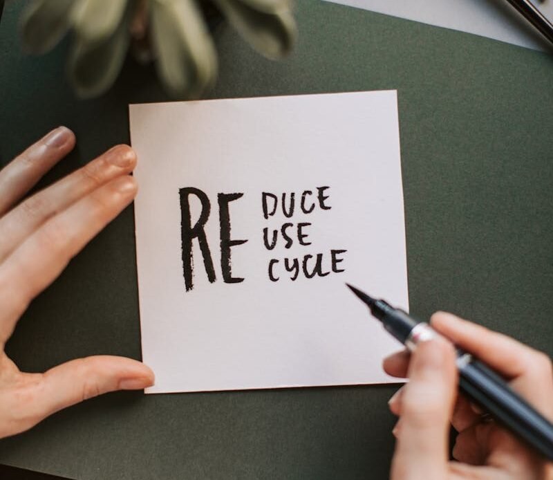 Close-up of hands writing a motivational 'Reduce Reuse Recycle' note on paper with a pen, surrounded by office items.